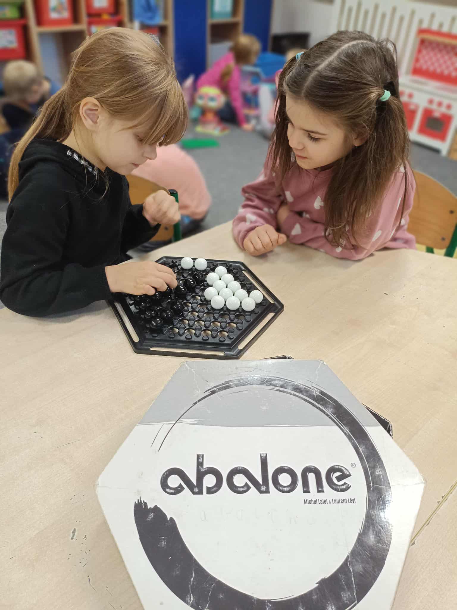 Dwie dziewczynki siedzą przy stole i skupiają się na rozgrywce w Abalone. Przed nimi stoi plansza oraz pudełko gry. W tle widać inne dzieci bawiące się w sali.