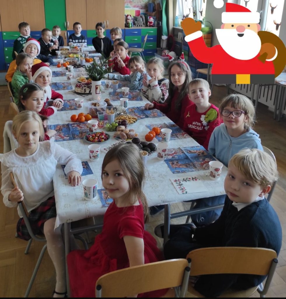 21 dzieci siedzi wokół nakrytego obrusem stołu, na którym są: wazon z gałązkami, słodkie i słone przekąski, owoce, serwetki, kubeczki. W prawym górnym rogu naklejka Mikołaja. W tle elementy sali lekcyjnej.