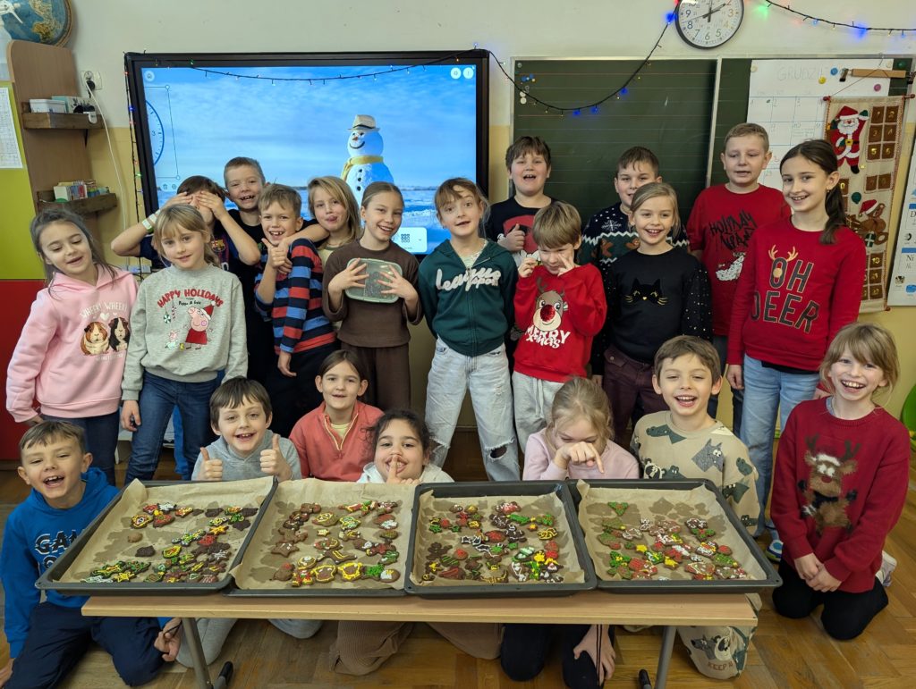 Grupa dzieci w sali lekcyjnej pozuje do wspólnego zdjęcia. Przed nimi na stole leżą blachy z kolorowymi, świątecznymi pierniczkami w różnych kształtach. W tle widać tablicę oraz ekran z zimową grafiką bałwana.