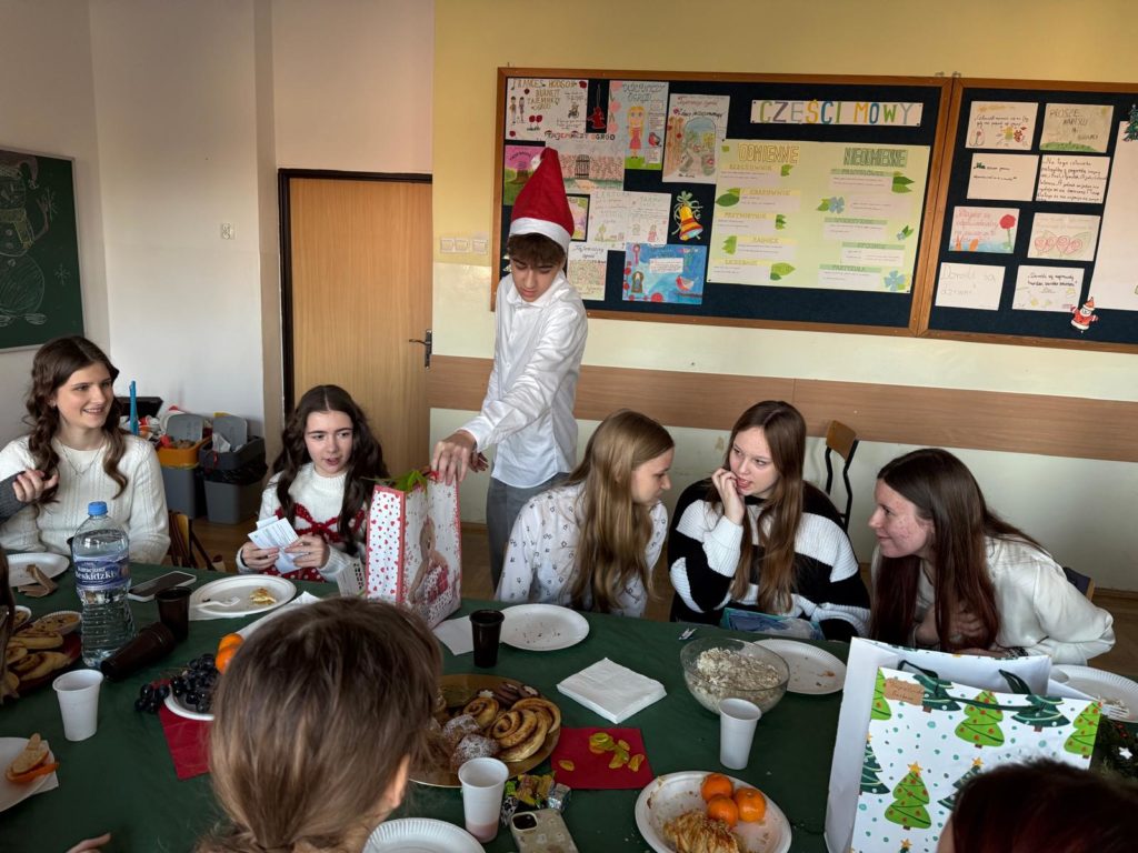 Uczniowie siedzą przy stole podczas klasowego spotkania świątecznego, a jeden z nich w czapce Mikołaja wręcza prezent; na stole leżą talerze z wypiekami, owoce i napoje, w tle gazetka szkolna.