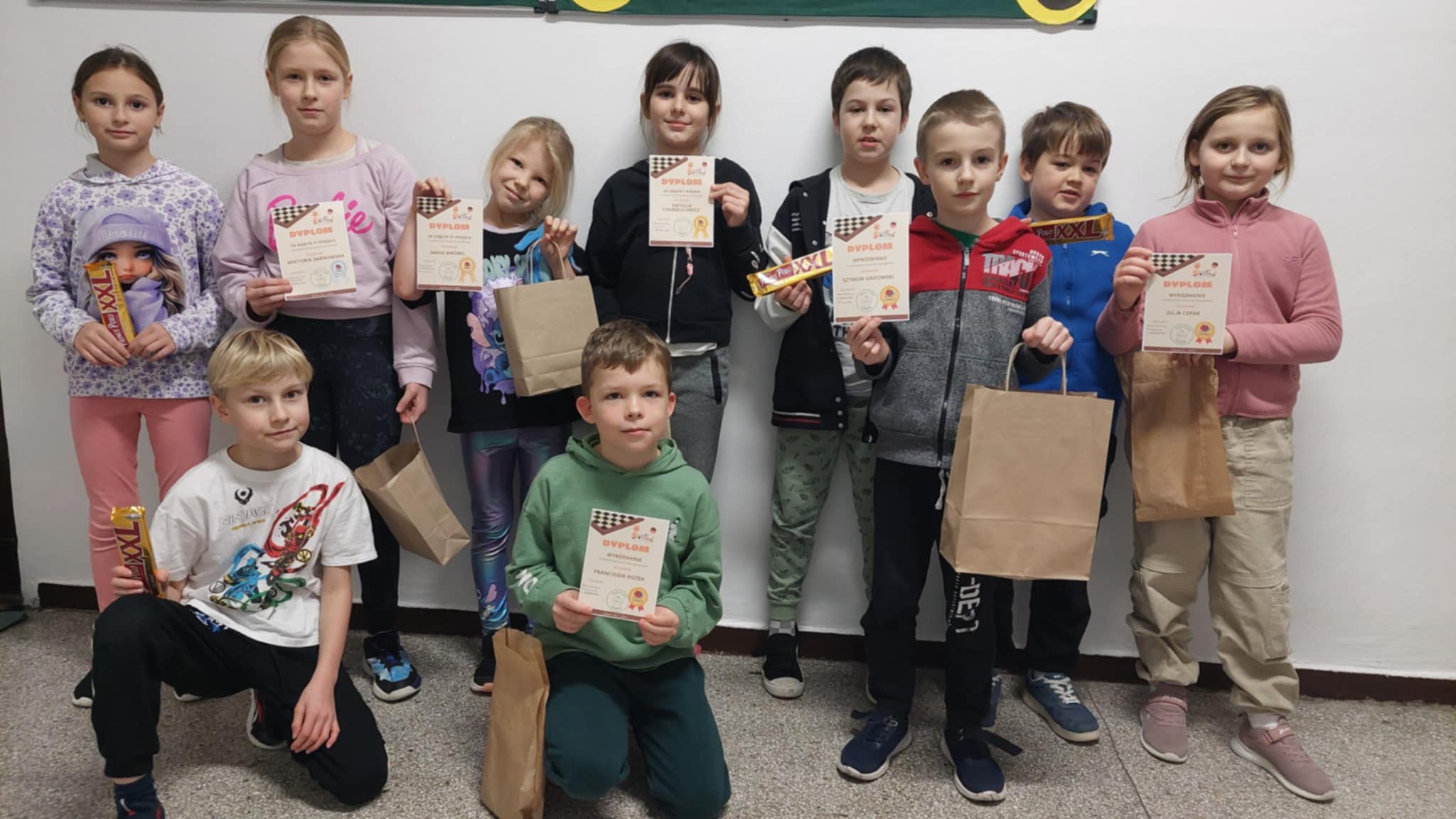 Grupa dzieci stoi razem pod ścianą i trzyma dyplomy oraz nagrody. Dzieci uśmiechają się i prezentują zdobyte wyróżnienia po turnieju warcabowym.