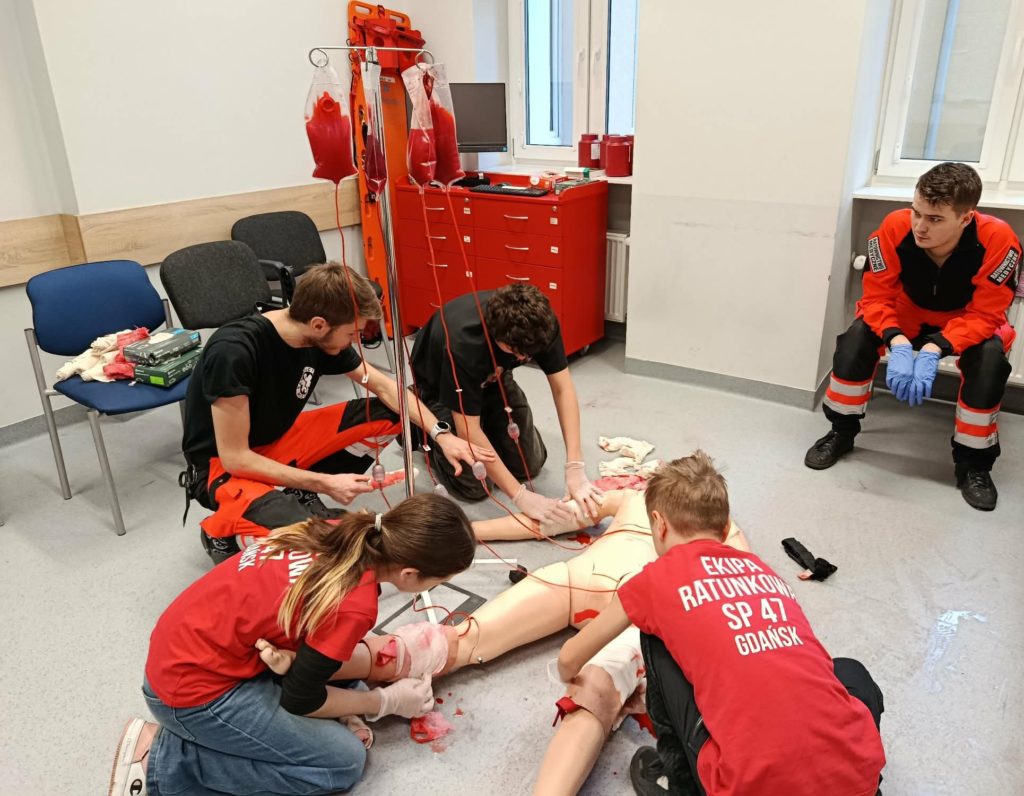 Na pierwszym planie leży fantom pozorujący krwotok z kończyn. Przy nim trzej uczniowie tamujący krwotoki i dwa studenci przyglądający się akcji