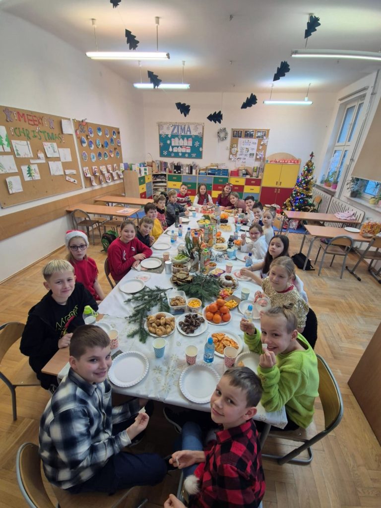 Dzieci siedzą przy prostokątnym stole .Na stole znajduje się jedzenie. Pomarańcze ciasta ,słone przekąski, woda , soki. a dziećmi jest ściana na której są dwie tablice z gazetkami informacyjnymi. Pod tablicami są półki szkolne. Po prawej stronie są okna i widać dwa stoliki- ławki szkolne.