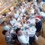 Uczniowie klasy 3c siedzą przy długim, świątecznie nakrytym stole w sali lekcyjnej. Na stole znajdują się owoce, słodki poczęstunek i napoje. Dzieci uśmiechają się do aparatu, w tle widoczne są kolorowe szafki i świąteczne dekoracje.