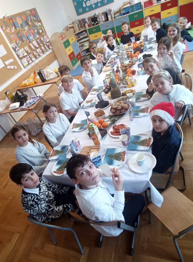 Uczniowie klasy 3c siedzą przy długim, świątecznie nakrytym stole w sali lekcyjnej. Na stole znajdują się owoce, słodki poczęstunek i napoje. Dzieci uśmiechają się do aparatu, w tle widoczne są kolorowe szafki i świąteczne dekoracje.