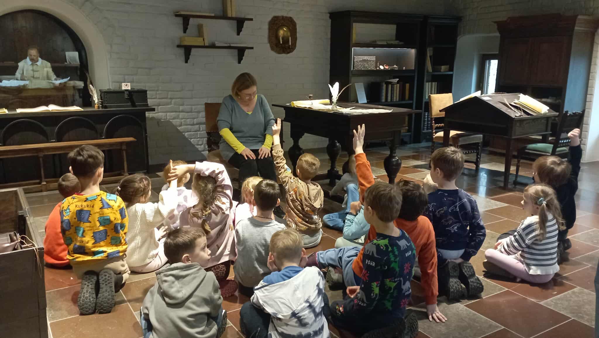 Na podłodze sali muzealnej siedzą w rozsypce uczniowie klasy 1c. Niektórzy podnoszą rękę do odpowiedzi. W tle zabytkowe meble. Na krześle siedzi kustoszka muzeum.