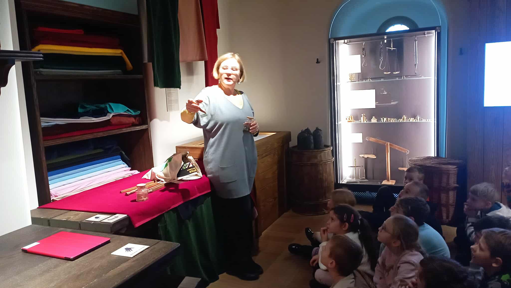 W sali muzealnej imitującej kantor kupca na tle eksponatów stoi kustoszka muzeum ze słoiczkiem przypraw korzennych w dłoni. W tle średniowieczne wagi i miary. Przy kustoszce siedzą w rozsypce uczniowie klasy 1c