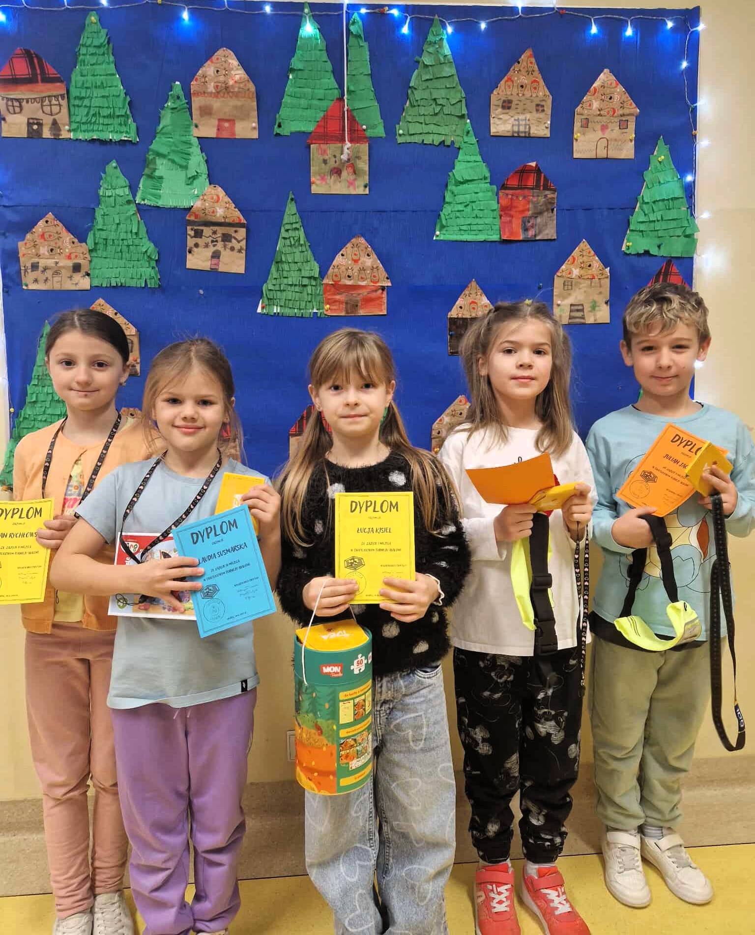 Grupa pięciorga dzieci stoi w szeregu i trzyma kolorowe dyplomy. W tle widać dekorację z papierowych choinek i domków oraz światełka. Dzieci uśmiechają się.
