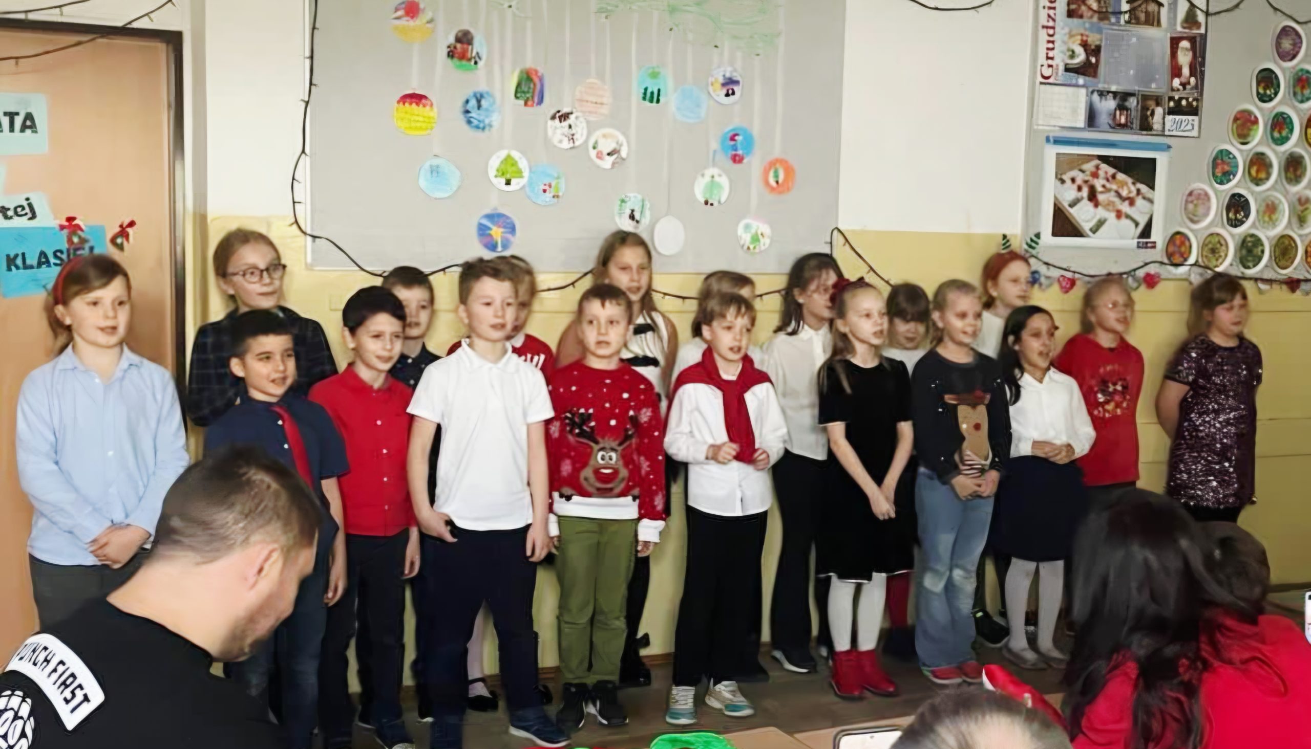 Grupa dzieci stoi pod ścianą w odświętnym ubraniu, nad nimi wiszą 2 tablice z świątecznymi dekoracjami.