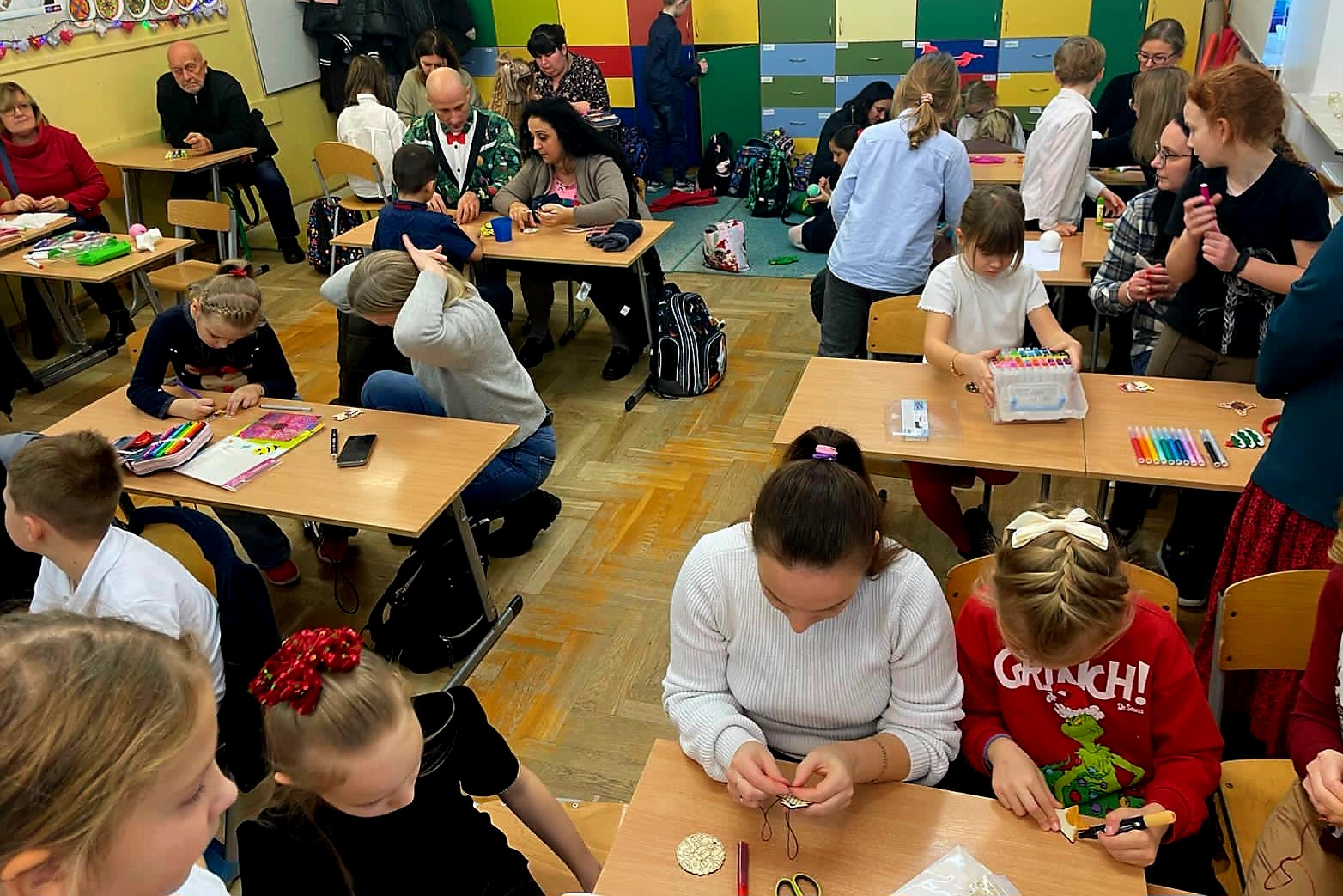 Rodzice wraz z dziećmi siedzą przy stołach, ozdabiając razem ozdoby bożonarodzeniowe.