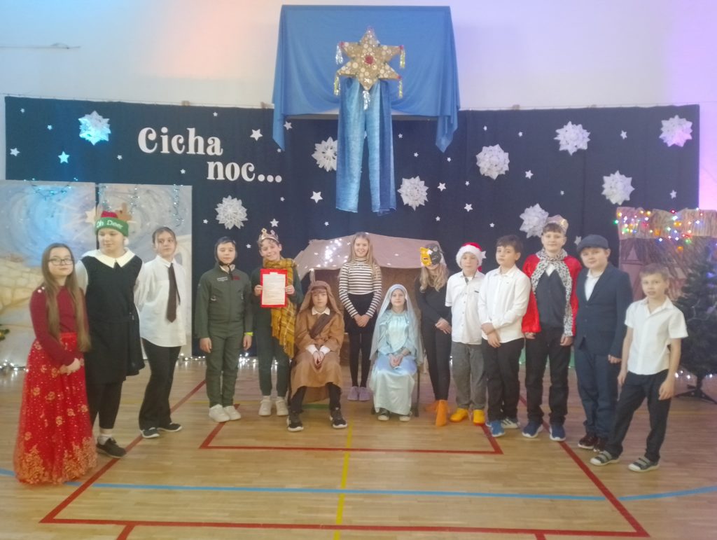 Na środku sceny dwoje dzieci siedzi przed stajenką na krzesłach. Są przebrani za Józefa i Maryję. Liczna grupa dzieci stoi w półkolu. Mają różne stroje, długie szaty, korony, chusty i ubrania codzienne. W tle gwiazda betlejemska, ciemne tło z gwiazdami , napis "Cicha noc" i świąteczne lampki