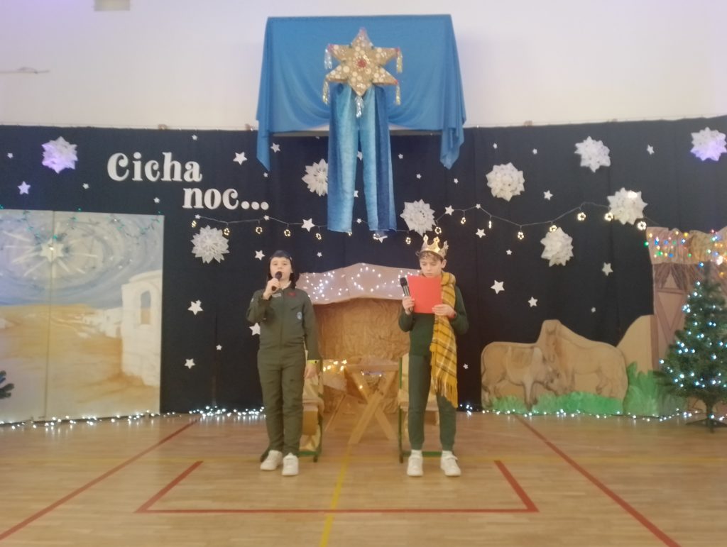 Na scenie dwóch chłopców. Po lewej stronie chłopiec ubrany w strój pilota. Trzyma mikrofon i mówi. Po prawej stronie chłopiec ubrany w koronę i owinięty dużym szalem trzyma czerwoną kartkę i czyta tekst. Za nimi stajenka, światełka i duża gwiazda betlejemska zawieszona wysoko. Czarne tło z gwiazdami i napisem "Cicha noc"