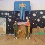 Na scenie dwóch chłopców. Po lewej stronie chłopiec ubrany w strój pilota. Trzyma mikrofon i mówi. Po prawej stronie chłopiec ubrany w koronę i owinięty dużym szalem trzyma czerwoną kartkę i czyta tekst. Za nimi stajenka, światełka i duża gwiazda betlejemska zawieszona wysoko. Czarne tło z gwiazdami i napisem "Cicha noc"