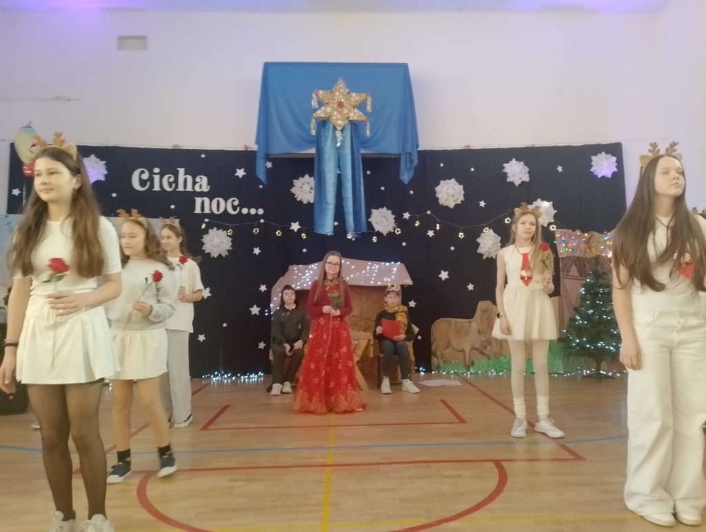 Po lewej i prawej stronie sceny stoją dziewczynki w jasnych strojach,mają opaski z rogami renifera i trzymają czerwone róże. Pośrodku sceny stoi dziewczynka w kolorowej,długiej sukni i trzyma w ręku różę. Z tyłu widać stajenkę i dwóch chłopców. Cała scena świątecznie udekorowana.