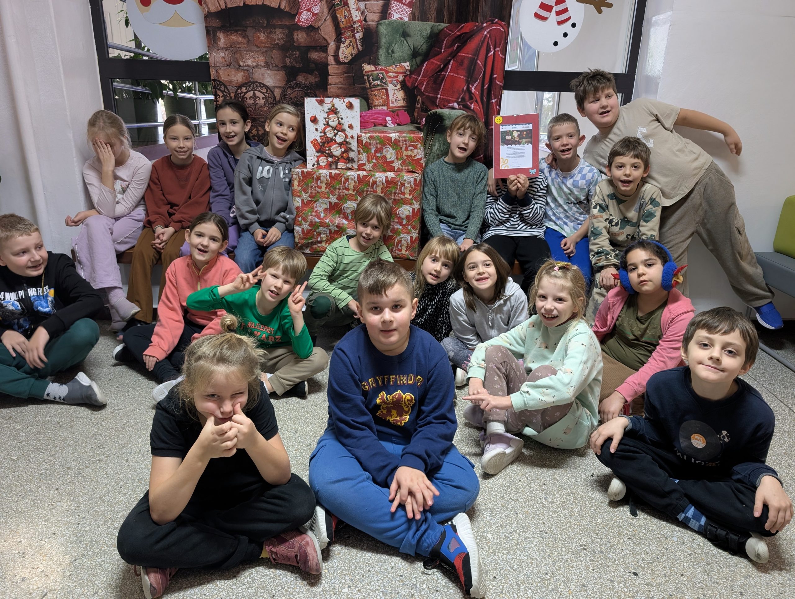 Grupa dzieci z klasy 2C siedzi i pozuje do zdjęcia na tle dużego stosu kolorowo zapakowanych prezentów świątecznych. Dzieci uśmiechają się i wyglądają na podekscytowane udziałem w akcji pomocowej. W tle znajduje się dekoracja z motywem kominka i Mikołaja, tworząca świąteczną atmosferę.