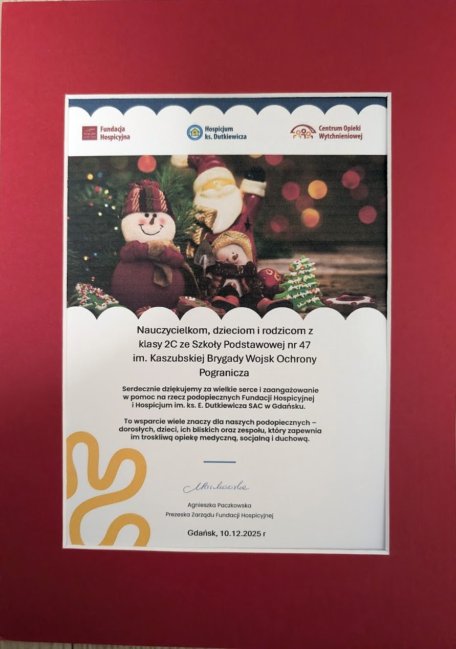 Zbliżenie na ozdobną ramkę z podziękowaniem od Fundacji Hospicyjnej dla nauczycieli, dzieci i rodziców z klasy 2C. Dokument umieszczony jest w czerwonej oprawie i zawiera świąteczną grafikę oraz tekst wyrażający wdzięczność za okazane serce i wsparcie podopiecznych hospicjum.