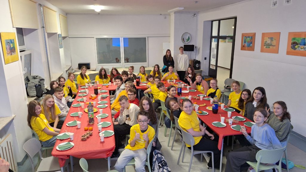 Grupa uczniów – wolontariuszy siedzących przy długich stołach, na których rozłożone są talerzyki. Na stole stoją napoje, przekąski i owoce.