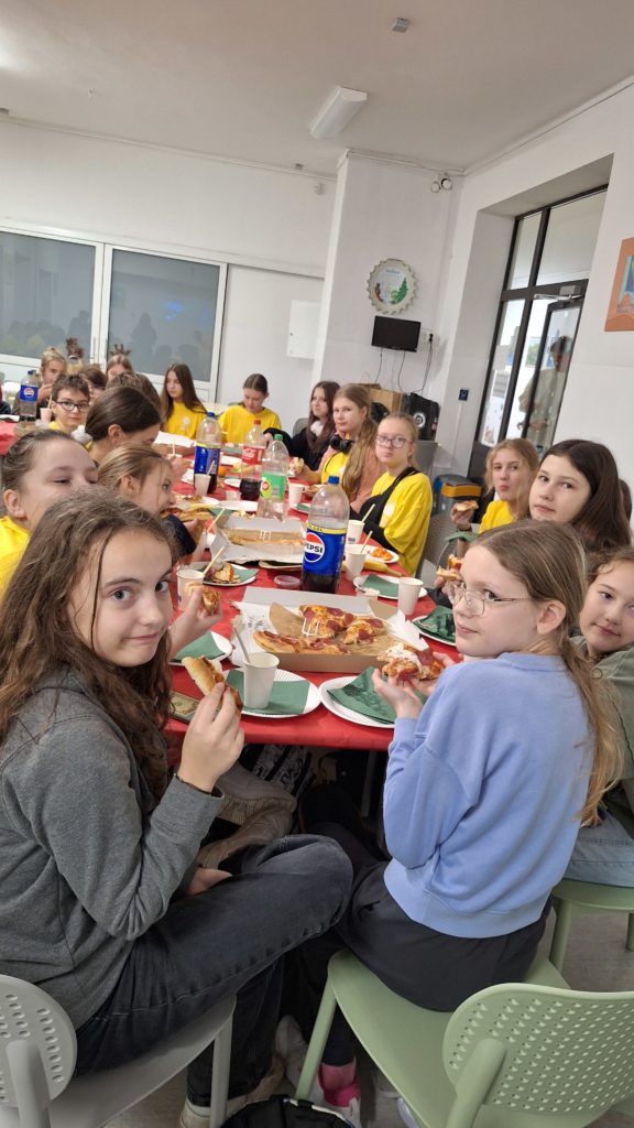 Grupa dzieci przy wspólnym stole jedzą pizzę i rozmawiają. Na stole leży pizza i stoją napoje.