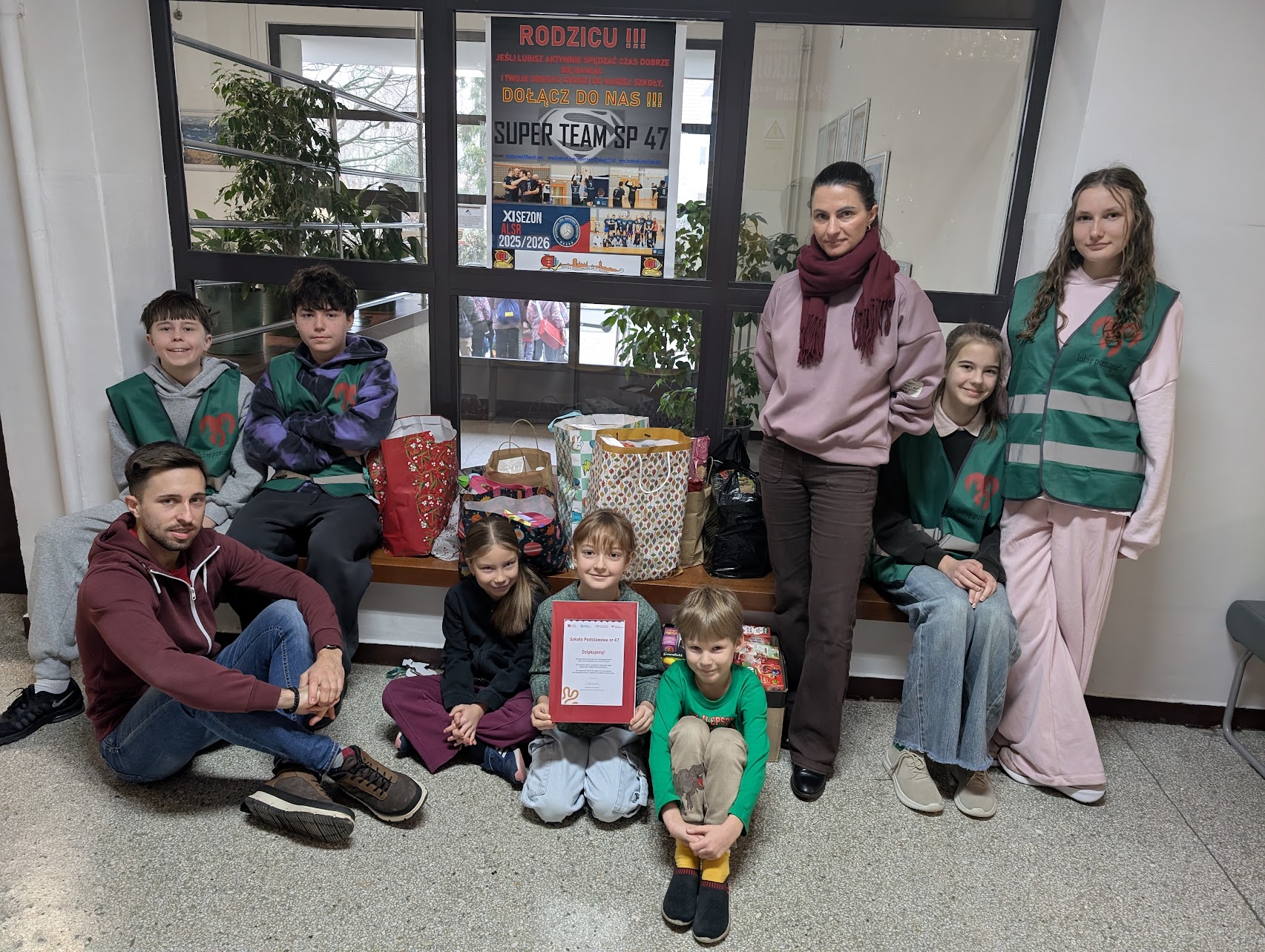 Grupa uczniów i nauczycieli siedzi i stoi wokół zebranych prezentów w szkolnym korytarzu. Przed nimi znajdują się kolorowe torby i paczki przygotowane w ramach zbiórki dla Fundacji Hospicyjnej. Dzieci i młodzież mają na sobie zielone kamizelki wolontariuszy. Wszyscy uśmiechają się do zdjęcia, a jedna z dziewczynek trzyma w rękach podziękowanie od fundacji. W tle widoczne są szkolne ogłoszenia i duże okno.