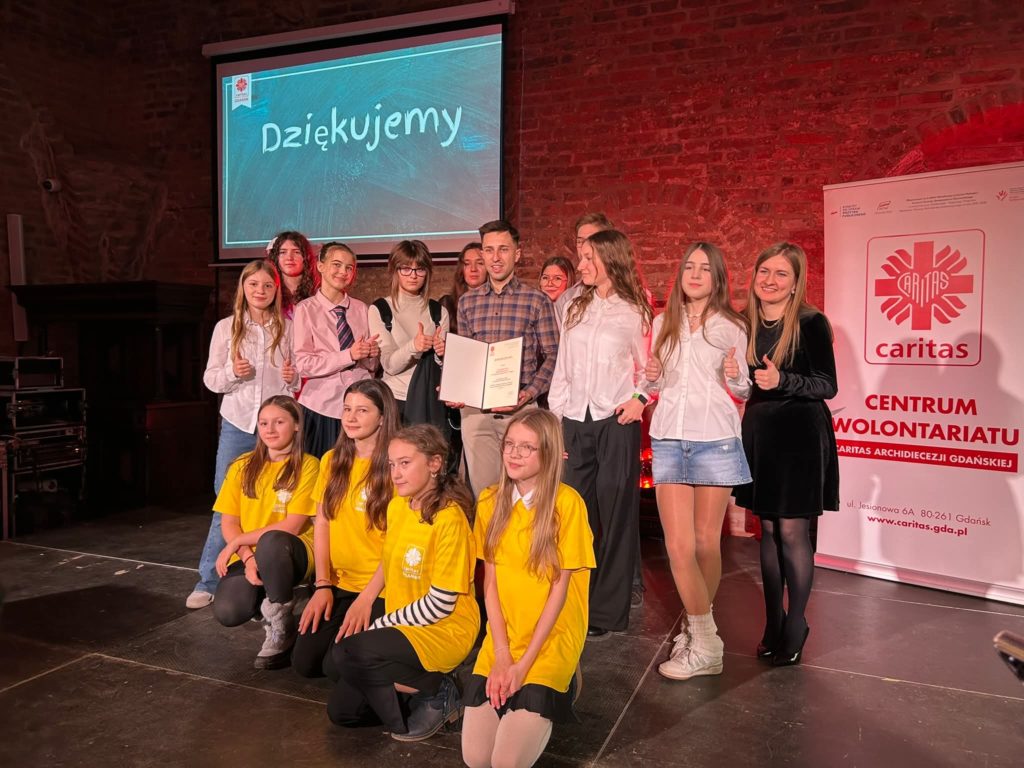 Grupa młodych wolontariuszek i wolontariuszy wraz z opiekunem stoi na scenie i pozuje do wspólnego zdjęcia. W centrum mężczyzna trzyma otwarty dyplom, a uczestnicy uśmiechają się i pokazują kciuki w górę. Część osób ubrana jest w białe koszule, kilka wolontariuszek z przodu ma żółte koszulki. W tle ekran z napisem „Dziękujemy” oraz baner Caritas Centrum Wolontariatu.