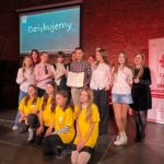 Grupa młodych wolontariuszek i wolontariuszy wraz z opiekunem stoi na scenie i pozuje do wspólnego zdjęcia. W centrum mężczyzna trzyma otwarty dyplom, a uczestnicy uśmiechają się i pokazują kciuki w górę. Część osób ubrana jest w białe koszule, kilka wolontariuszek z przodu ma żółte koszulki. W tle ekran z napisem „Dziękujemy” oraz baner Caritas Centrum Wolontariatu.