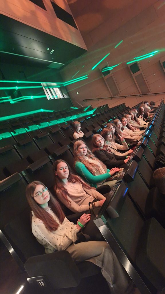 Grupa młodych osób siedzi w jednym długim rzędzie foteli w sali konferencyjnej. Za ich plecami kolejne rzędy pustych foteli. Ich ubiór wskazuje na eleganckie spotkanie.