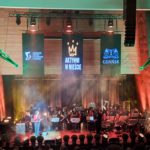 Scena z wydarzenia, podczas którego występuje orkiestra. W tle zawieszone są trzy duże banery: z logo Gdańska, Regionalnego Centrum Wolontariatu w Gdańsku oraz hasłem „Aktywni w mieście”. Scena jest dynamicznie oświetlona i ozdobiona roślinami w donicach. Na scenie jest prowadząca oraz dyrygent orkiestry. Po lewej stronie sceny znajduje się charakterystyczny element graficzny w kształcie dźwigu, nawiązujący do symbolu Gdańska.