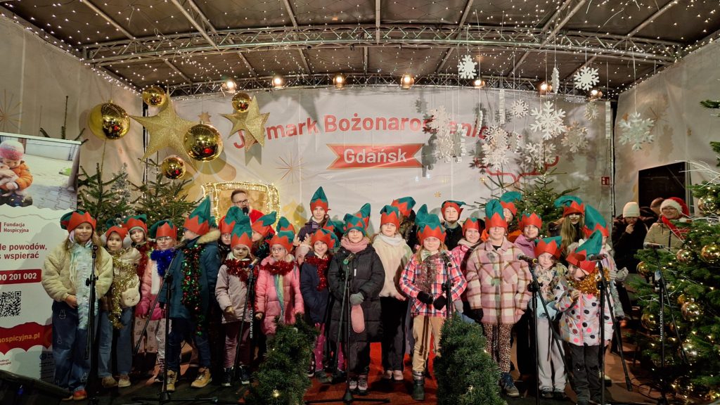 Dziecięcy zespół szkolny „Kropka” stoi na scenie podczas Jarmarku Bożonarodzeniowego w Gdańsku. Uczniowie ubrani są w zimowe kurtki i czapki elfów, przed nimi ustawione są mikrofony. W tle widać świąteczne dekoracje, choinki, lampki oraz baner wydarzenia. Scena ma radosny, świąteczny charakter, a występ odbywa się w ramach koncertu charytatywnego na rzecz hospicjum.