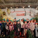 Dziecięcy zespół szkolny „Kropka” stoi na scenie podczas Jarmarku Bożonarodzeniowego w Gdańsku. Uczniowie ubrani są w zimowe kurtki i czapki elfów, przed nimi ustawione są mikrofony. W tle widać świąteczne dekoracje, choinki, lampki oraz baner wydarzenia. Scena ma radosny, świąteczny charakter, a występ odbywa się w ramach koncertu charytatywnego na rzecz hospicjum.