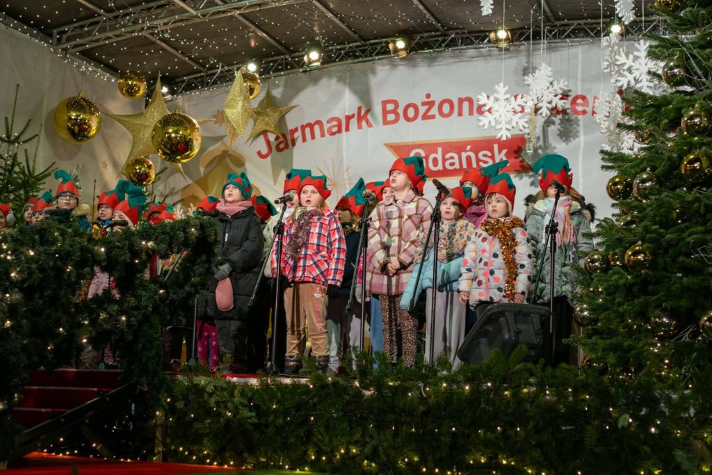 Dziecięcy zespół szkolny „Kropka” stoi na scenie podczas Jarmarku Bożonarodzeniowego w Gdańsku. Uczniowie ubrani są w zimowe kurtki i czapki elfów, przed nimi ustawione są mikrofony. W tle widać świąteczne dekoracje, choinki, lampki oraz baner wydarzenia. Scena ma radosny, świąteczny charakter, a występ odbywa się w ramach koncertu charytatywnego na rzecz hospicjum.