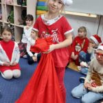 Dziewczynka w czerwonym ubraniu i czapce mikołaja stoi w sali świetlicowej i trzyma wielki worek z prezentami, w koło niej siedzą na podłodze inne dzieci, każde z nich ma ubrane na sobie świąteczne akcenty