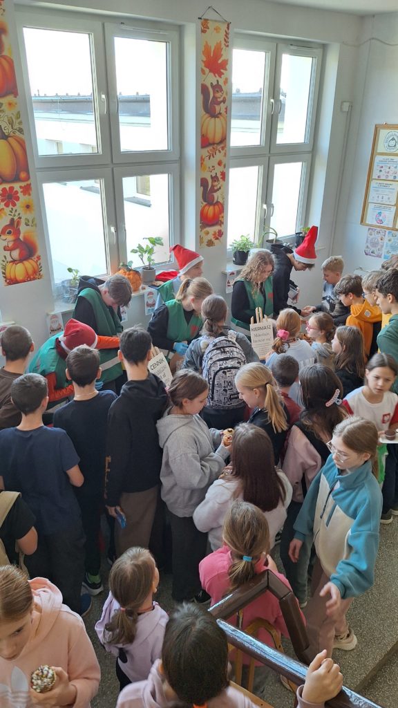 Tłum uczniów gromadzi się na szkolnym korytarzu wokół stoiska kiermaszu. Kilkoro dzieci w kamizelkach wolontariuszu i czapkach Mikołaja sprzedaje wypieki.