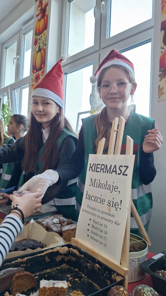 Dwie dziewczynki w czapkach Mikołaja i kamizelkach wolontariuszy stoją za stołem z domowymi ciastami. Jedna z nich podaje wypiek kupującej osobie. Obok nich znajduje się tabliczka z informacją o charytatywnym kiermaszu oraz o tym, że dochód przeznaczony jest na prezenty dla kilku dzieci.