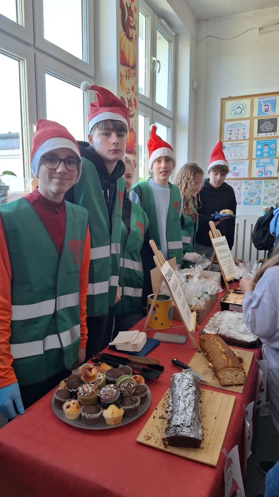 Grupa uczniów w kamizelkach wolontariuszy i czapkach Mikołaja stoi za stołem kiermaszu szkolnego. Na stole leżą babeczki, ciasta. Dzieci uśmiechają się i obsługują kupujących na korytarzu szkolnym.