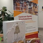 Na zdjęciu jest stojak z banerem Fundacji Hospicyjnej i Centrum Opieki Wytchnieniowej, a na stoliku przed nim leżą zapakowane świąteczne prezenty przewiązane wstążkami.