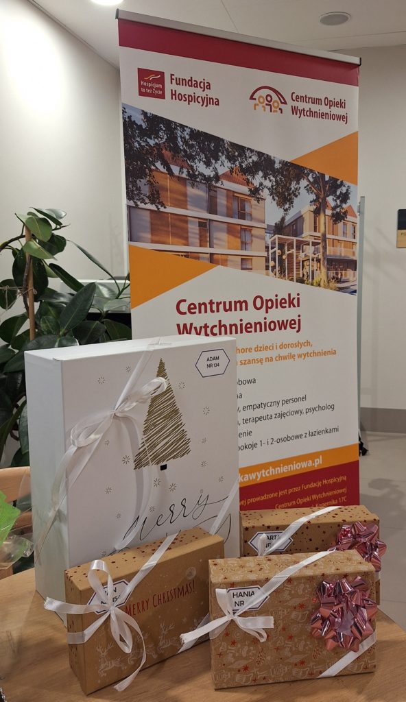 Na zdjęciu jest stojak z banerem Fundacji Hospicyjnej i Centrum Opieki Wytchnieniowej, a na stoliku przed nim leżą zapakowane świąteczne prezenty przewiązane wstążkami.