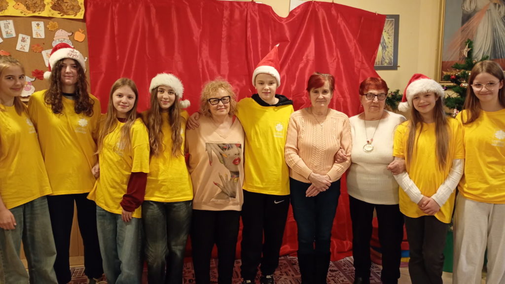 Grupa wolontariuszy w koszulkach wolontariuszy stoi w szeregu. Między nimi są trzy starsze kobiety. Na głowach niektórych czapka Mikołaja. Wszyscy trzymają się za ramiona.