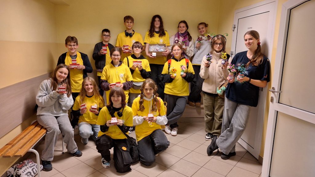 Grupa wolontariuszy stoi na szkolnym korytarzu. Część z nich kuca. Wszyscy trzymają w ręku małe pudełeczka z podarkami. Na ich twarzach jest uśmiech.