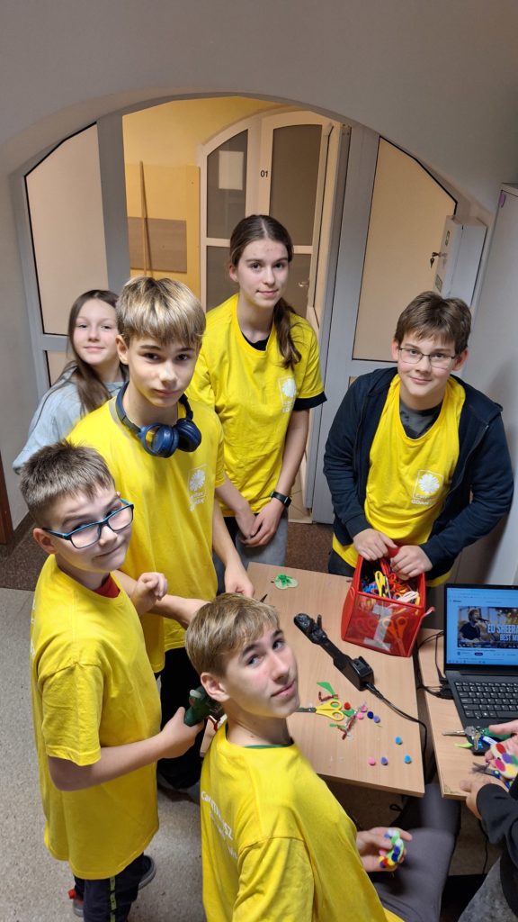 Grupa wolontariuszy w koszulkach wolontariuszy stoją przy małym stoliku, na którym są elementy dekoracyjne i pudełko z nożyczkami. Jedna osoba siedzi na krześle. Wszyscy patrzą w stronę aparatu. 