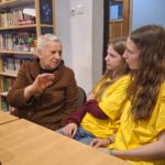 Starsza kobieta mówi do dwóch dziewczyn w koszulkach wolontariusza. Pokazuje coś ręką jakby coś tłumaczyła. Siedzą przy stole.