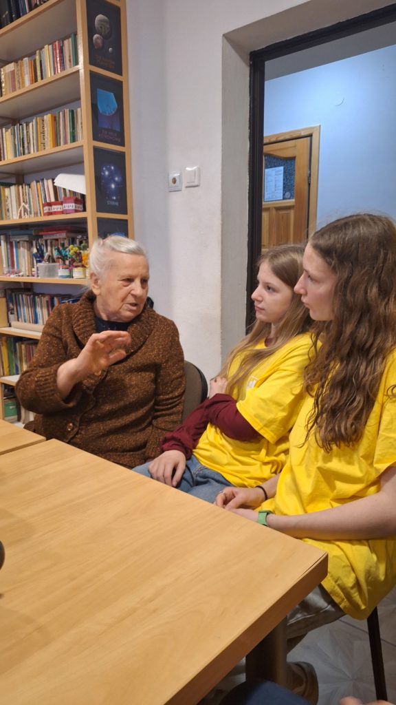 Starsza kobieta mówi do dwóch dziewczyn w koszulkach wolontariusza. Pokazuje coś ręką jakby coś tłumaczyła. Siedzą przy stole.