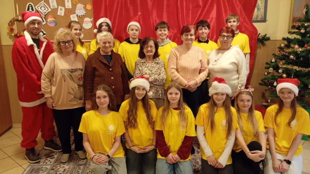 Grupa wolontariuszy oraz seniorów. Wolontariusze ubrani w koszulki wolontariuszy oraz w czapki Mikołaja. Jest też Mikołaj w stroju. W tle choinka ozdobioną świątecznymi dekoracjami, a na twarzach wszystkich jest uśmiechy.