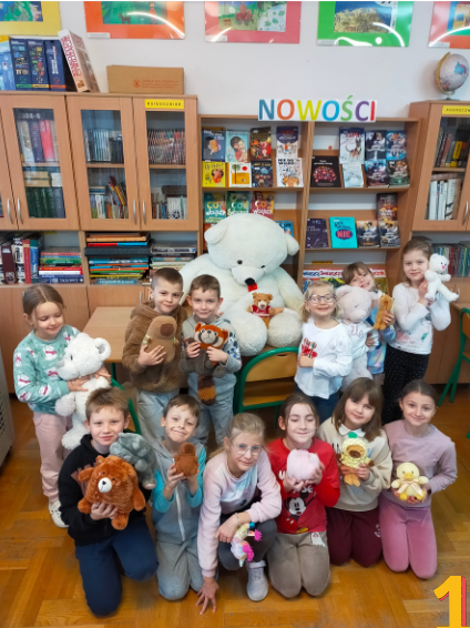 W prawym dolnym rogu zdjęcia cyfra 1. Czternaścioro dzieci pozuje w szkolnej bibliotece z pluszowymi zabawkami. W centrum stoi jeden bardzo duży biały miś siedzący na krześle. W tle widoczne są regały z książkami oraz kolorowe prace plastyczne.”