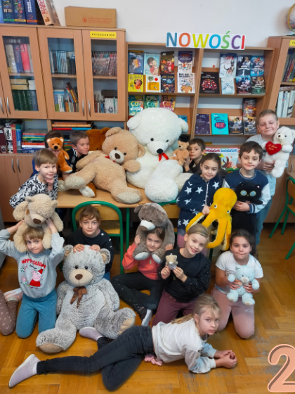W prawym dolnym rogu zdjęcia cyfra 2. Trzynaścioro dzieci stoi w szkolnej bibliotece, trzymając swoje pluszowe zabawki. Na stole przed nimi znajduje się jeden ogromny biały miś. W tle widać regały z książkami i prace plastyczne zawieszone na ścianie.”