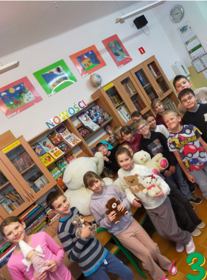 W prawym dolnym rogu zdjęcia cyfra 3. Siedmioro dzieci stoi w rzędzie w bibliotece szkolnej, każde trzyma pluszowego misia. Obok nich, na krześle, siedzi jeden duży biały miś. Za dziećmi widoczne są półki z książkami oraz kolorowe ilustracje na ścianie.”