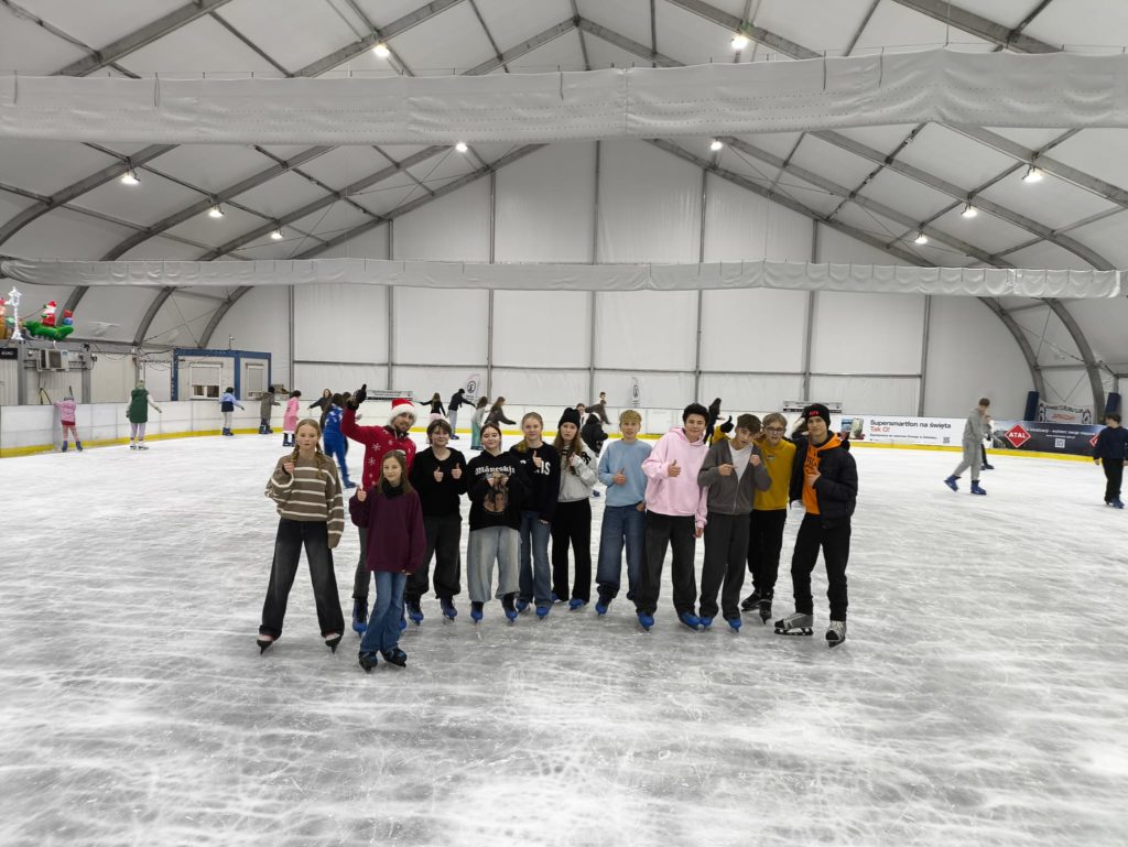 Grupa młodych wolontariuszy, którzy wspólnie spędzają czas na lodowisku. Wszyscy uśmiechają się i pozują do zdjęcia podnosząc kciuk do góry. W tle inni uczestnicy, którzy cieszą się jazdą na łyżwach.