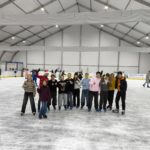 Grupa młodych wolontariuszy, którzy wspólnie spędzają czas na lodowisku. Wszyscy uśmiechają się i pozują do zdjęcia podnosząc kciuk do góry. W tle inni uczestnicy, którzy cieszą się jazdą na łyżwach.