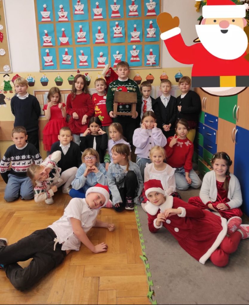 21 dzieci ustawionych w dwa rzędy, jeden stoi, drugi siedzi, dwoje uczniów leży z przodu w czapkach Mikołaja. Chłopiec stojący z tyłu trzyma w dłoniach karmnik dla ptaków. W prawym górnym roku naklejka Mikołaja. W tle ściana oraz tablica z pracami plastycznymi - Mikołajami.