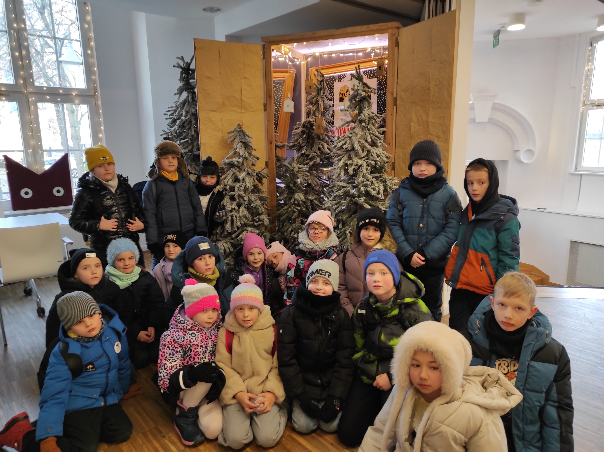 20 dzieci ustawionych w 2 rzędy, jeden stoi, drugi kuca. W tle witryna z otwartymi drzwiczkami, a w niej plakat przedstawienia "Opowieść wigilijna". Przed witryną ustawione 3 ośnieżone iglaki. Z lewej strony okno i logo Miejskiego Teatru Miniatura, z prawej fragment ściany.