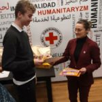 W sali konferencyjnej Pomorskiego Oddziału Okręgowego PCK na tle baneru PCK stoi uśmiechnięty uczeń, któremu organizatorka Olimpiady Zdrowia wręcza statuetkę jabłka, dyplom oraz nagrodę za zajęcie drugiego miejsca.